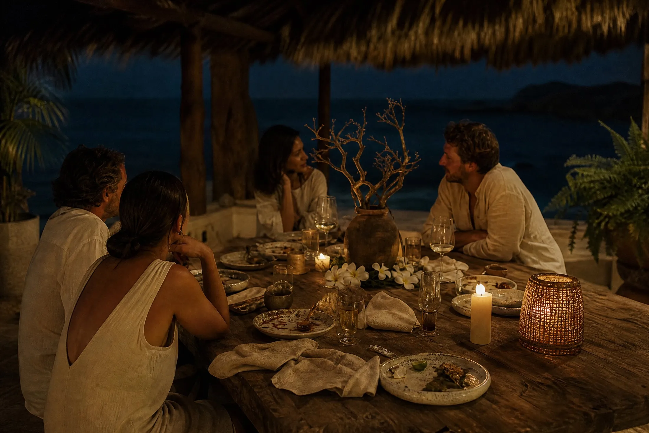 A candlelit dinner still going on a long carved teak table under palapa thatch, dark ocean behind